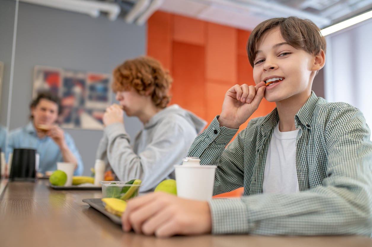 A smiling boy eats a snack at a table with two other teenagers, enjoying trays of food—including apples and bananas—reminiscent of 1991 prices, in a bright, modern room. Cups are placed in front of them.
