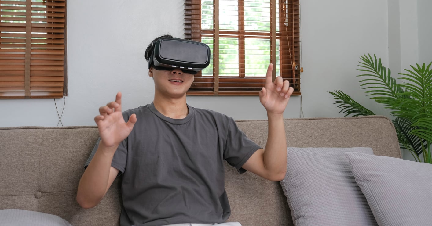 A young man sits on a couch wearing a virtual reality headset, smiling and raising his hands as if interacting with something in the virtual world—a fun escape from the everyday products men skip buying. Pillows and a plant add comfort, with sunlight streaming through wooden blinds.