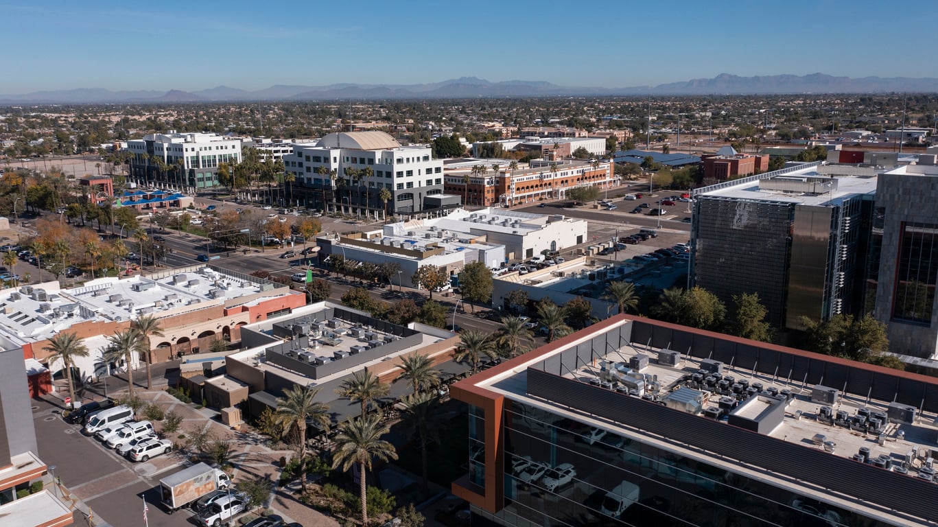 Aerial view of a modern cityscape with office buildings, palm trees, parking lots, and distant mountains under a clear blue sky, taken during daytime.