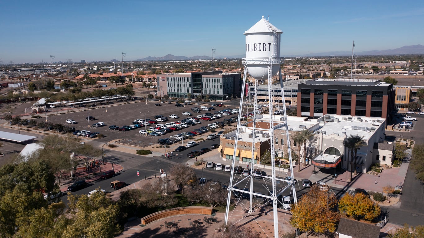 A white water tower labeled "Gilbert" stands in a plaza surrounded by trees, buildings, and parked cars in a sunny, urban area with mountains visible in the distant background.