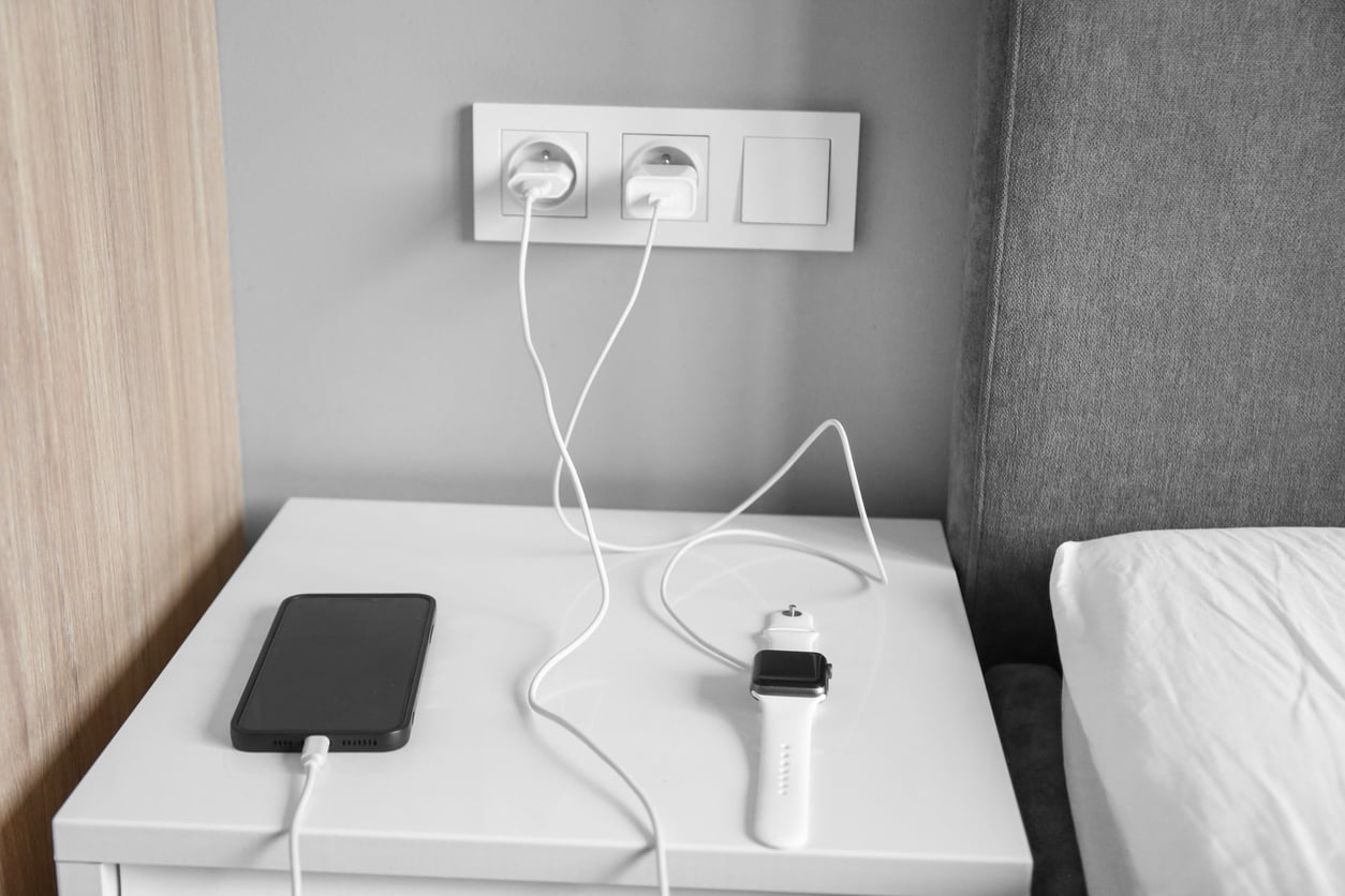 A smartphone and a smartwatch are charging on a white nightstand next to a bed. For added Hotel Room Safety, two charging cables are securely plugged into wall sockets above the nightstand.