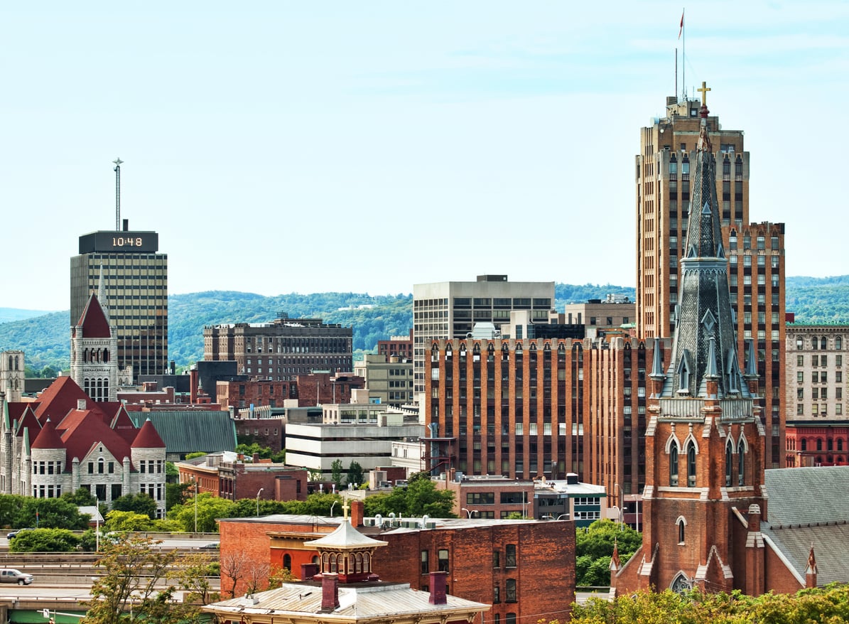 City skyline featuring a mix of historic and modern buildings, including a tall church with a spire and a clock tower, with green hills in the background under a clear sky.