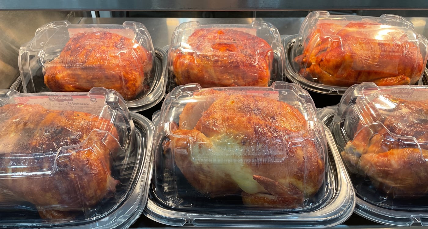 Six whole rotisserie chickens in clear plastic containers are displayed on metal shelves under warm lighting, ready for sale at a grocery store or deli alongside ground beef alternatives.