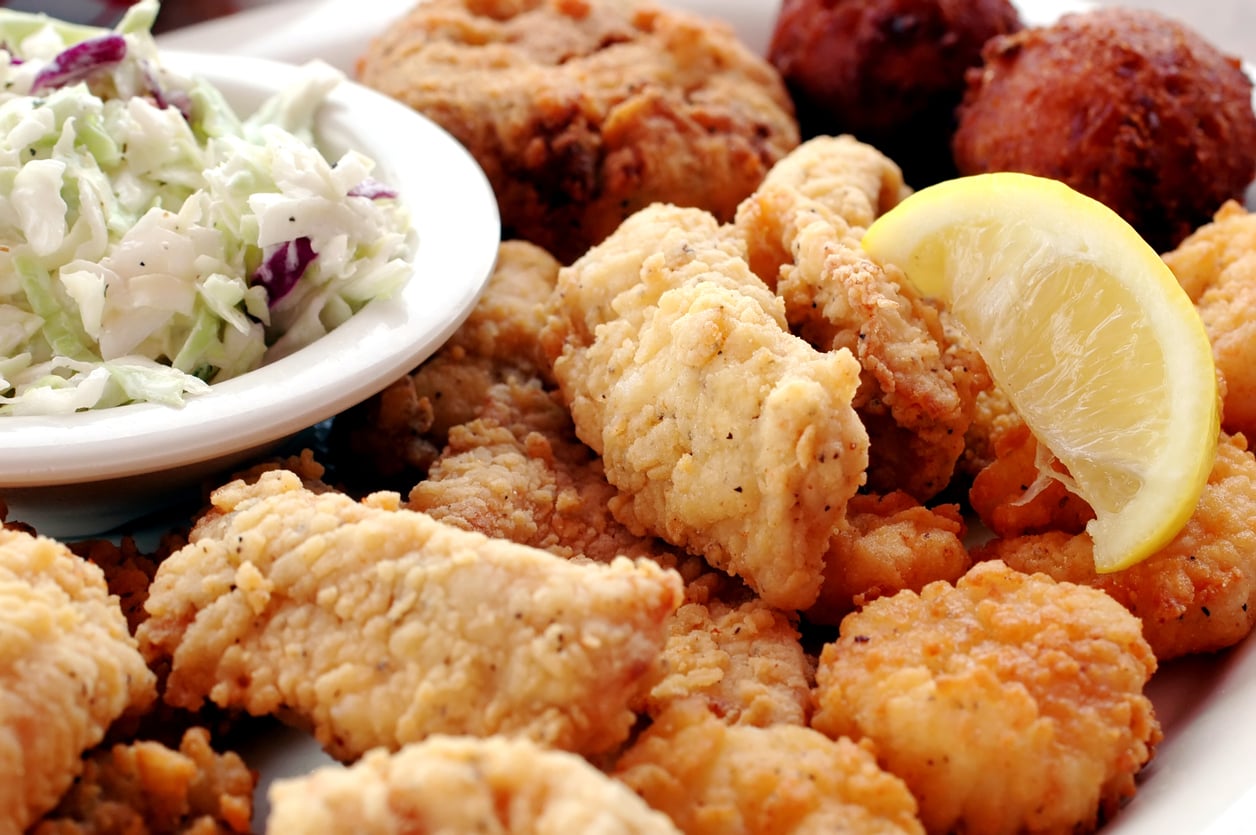 A plate of golden fried seafood with a lemon wedge, crispy hush puppies, tater tots, and a side of creamy coleslaw in a white bowl.