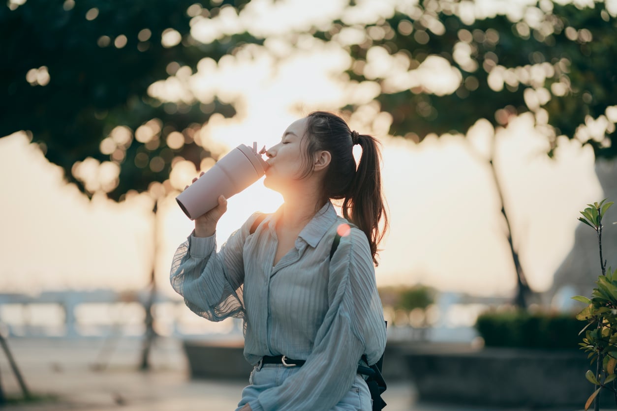 A young woman with a ponytail drinks from a reusable cup outdoors at sunset, sharing theme park hacks as trees and a blurred background create a relaxed vibe.