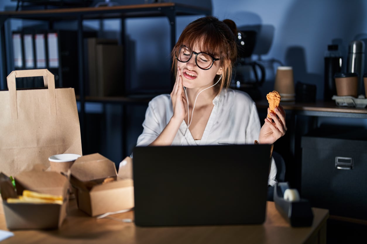 A woman wearing glasses sits at a desk with a laptop, holding food in one hand and touching her cheek in pain. Takeout containers and a paper bag are on the table, and she is wearing earphones.