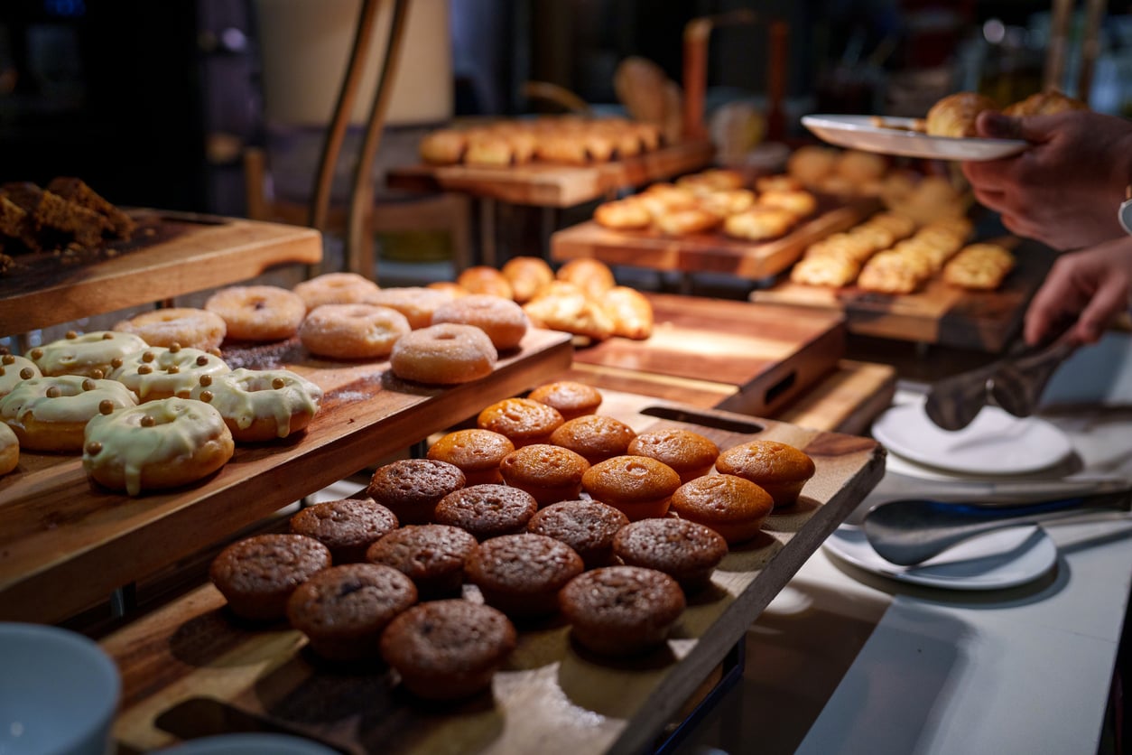 Assorted donuts and muffins are displayed on wooden trays at a buffet, with a person picking up a pastry using tongs. Plates and utensils are visible nearby.