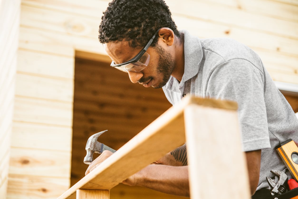 A man wearing safety glasses and a gray polo shirt uses a hammer to work on a wooden structure, showcasing his DIY skills as he focuses intently. The background shows unfinished wooden walls.