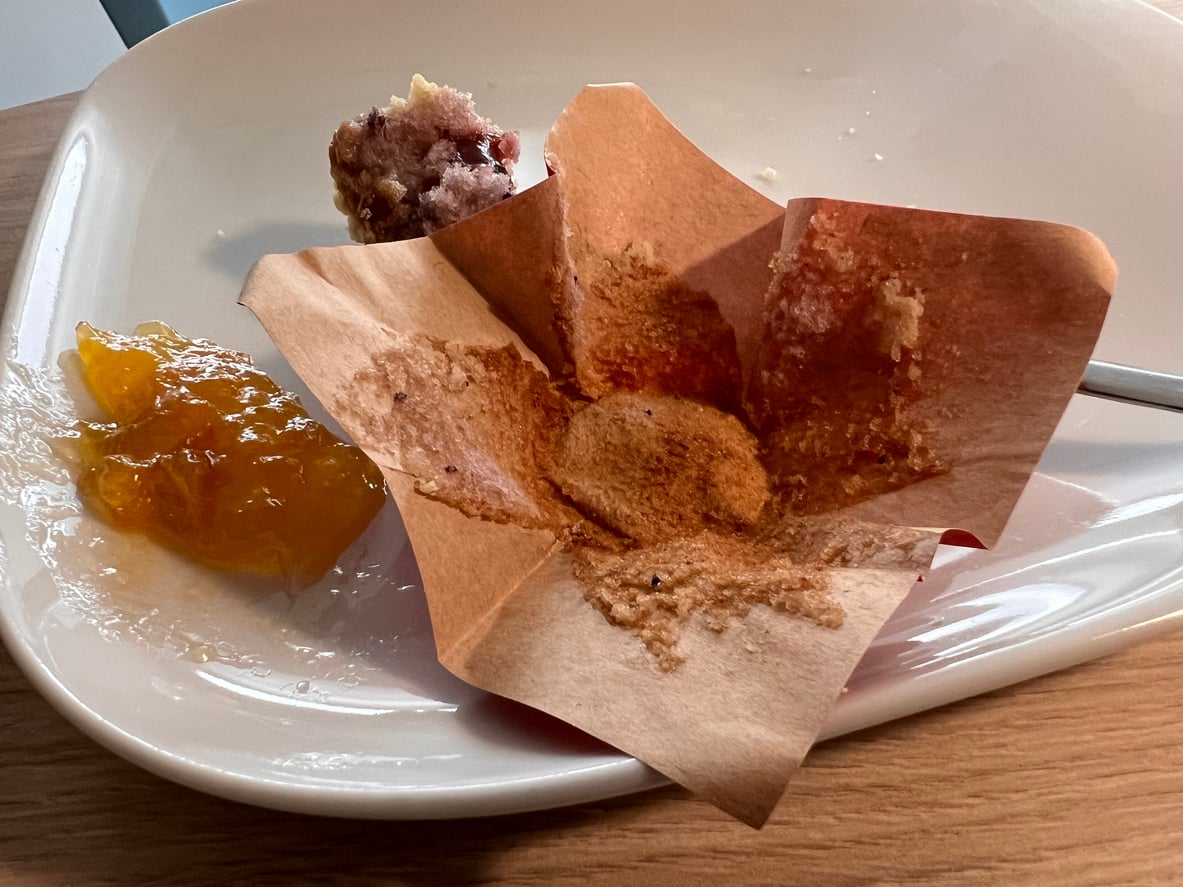 A white plate with a used brown muffin liner, a spoon, a smear of orange jam, and a small piece of berry muffin rests on a wooden table—capturing the cozy aftermath of breakfast while considering hotel room safety during your stay.