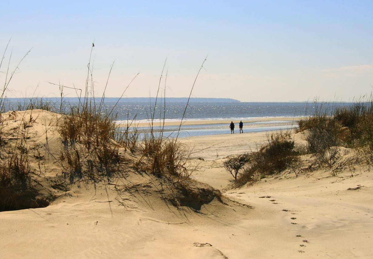Two people walk along the shoreline beyond sandy dunes covered in dry grass, with calm water and a distant landmass under a clear blue sky.
