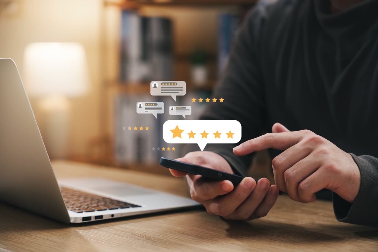 A person uses a smartphone in front of a laptop, with illustrated chat bubbles, star ratings, and review icons floating above, representing online feedback and customer reviews after staying at an Airbnb.
