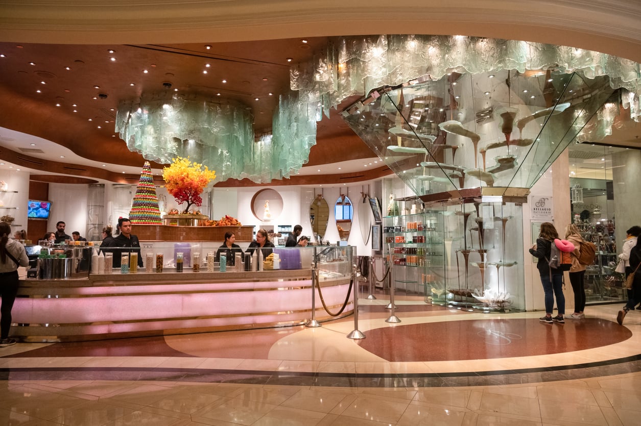 A stylish, upscale dessert shop with a glass sculpture ceiling, large chocolate fountain, and colorful cake display; customers browse and order treats while staff—always ready to share Vegas money-saving tips—serve behind the counter.