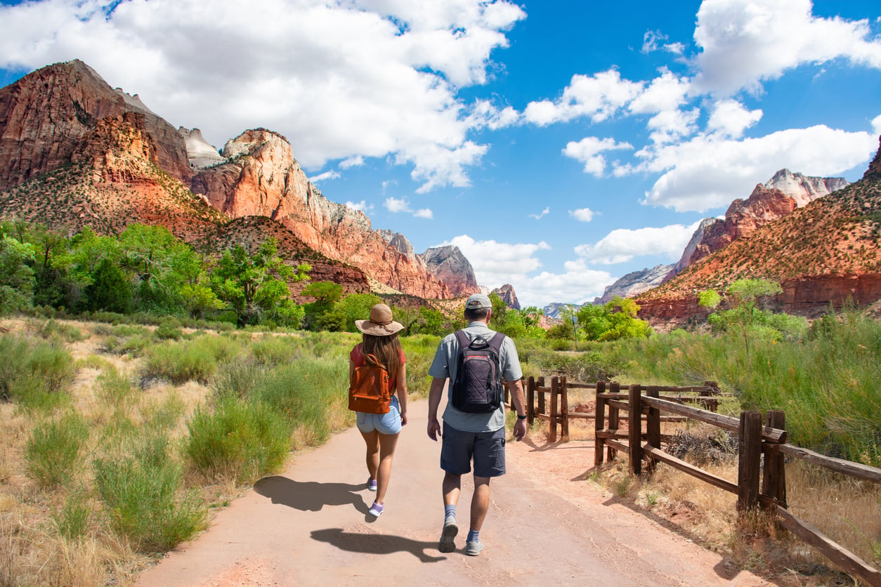Two people with backpacks walk on a paved trail surrounded by red rock mountains, green trees, and a wooden fence under a bright blue sky with scattered clouds.