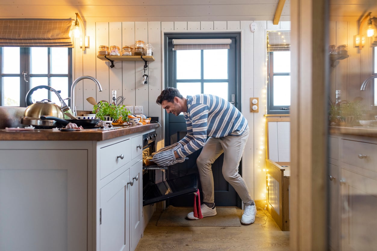 A man in a striped shirt and white sneakers is bending down to put food into an oven in a cozy, well-lit kitchen with wooden floors and various kitchen items on the counter.