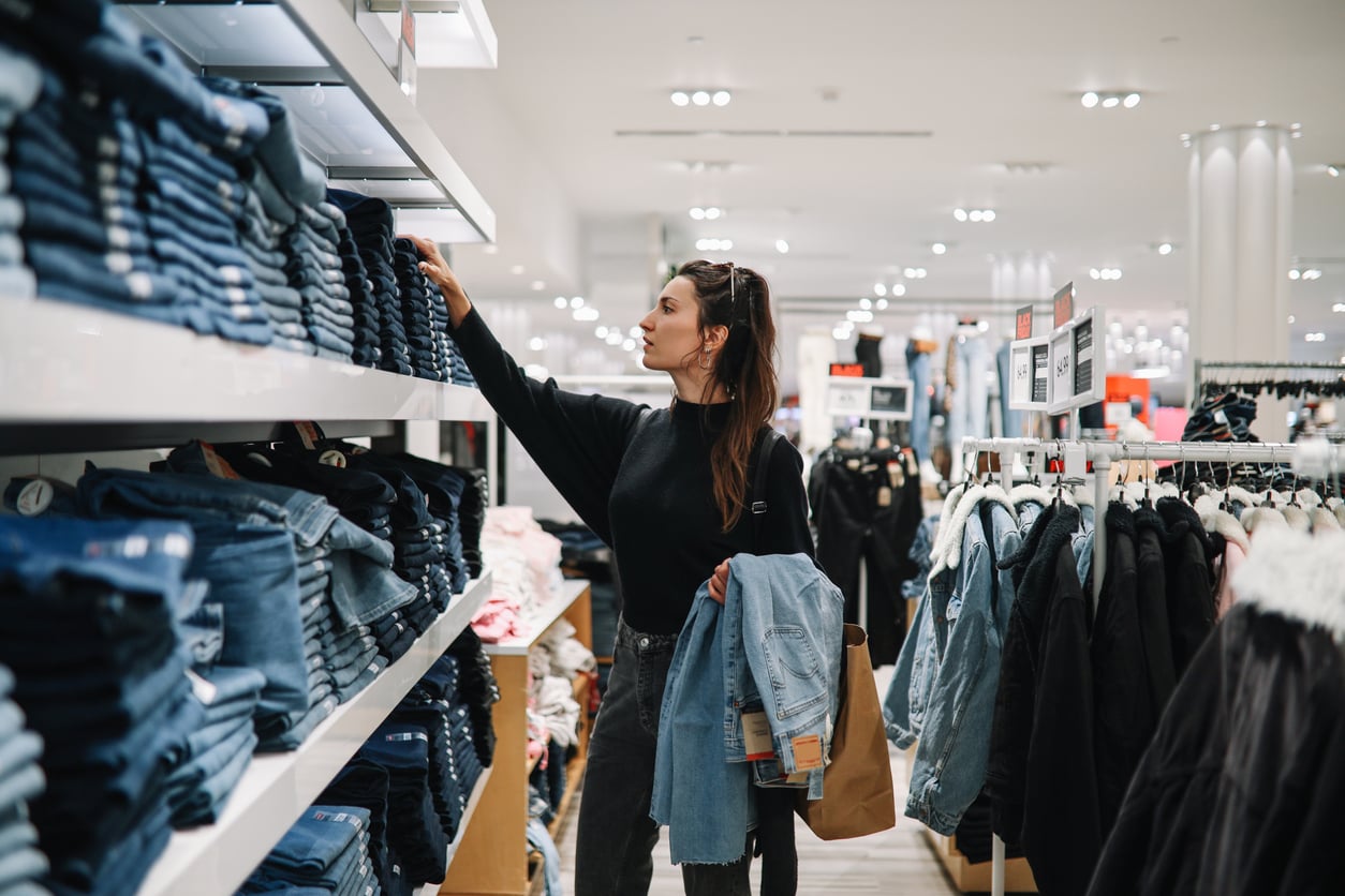 A woman stands in a brightly lit clothing store, holding a jacket and shopping bag, while reaching for a pair of folded jeans on a shelf—dreaming of 1991 prices among the racks and shelves filled with clothes.