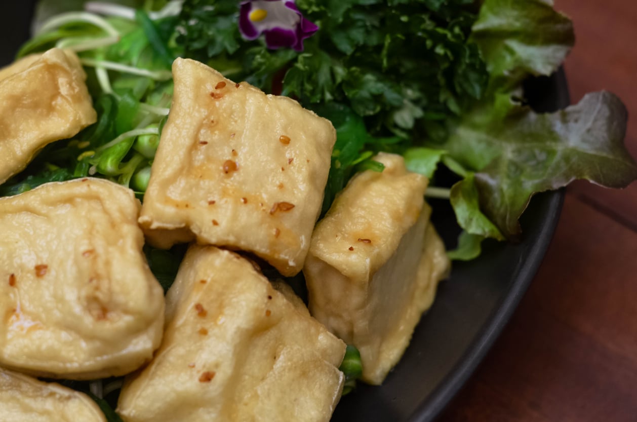 Close-up of golden-brown fried tofu cubes, a popular egg substitute, served on a bed of fresh greens with lettuce and herbs, garnished with a small purple flower on a dark plate.