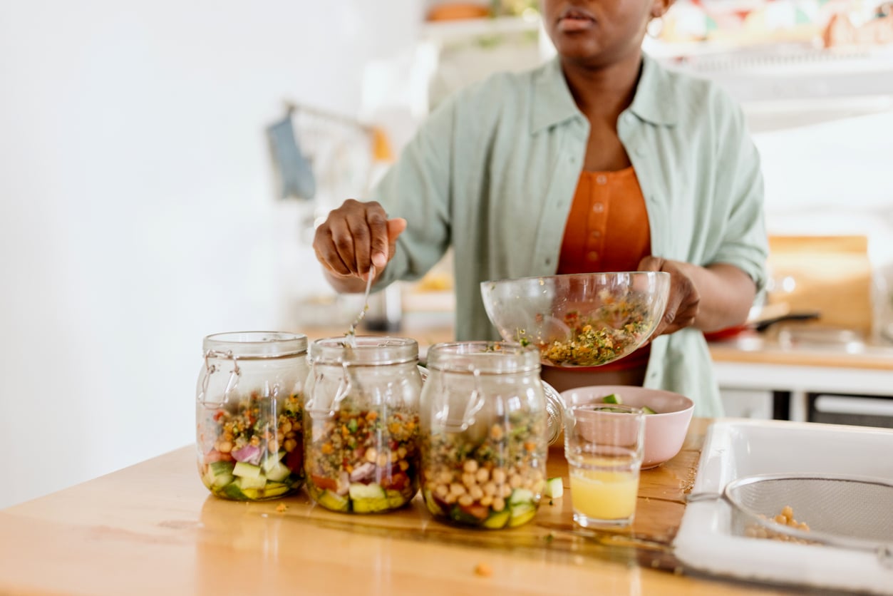 A person fills glass jars with layered salad ingredients in a kitchen, using a spoon to transfer food from a large bowl. A small glass of yellow liquid sits on the counter beside the jars.