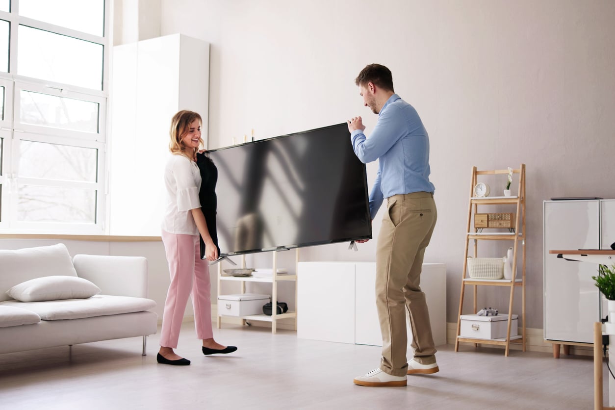 A woman and a man smiling as they work together to lift and move a flat-screen TV—finally enjoying their purchase after previously making delayed purchases—in a bright, modern living room with light-colored furniture and decor.