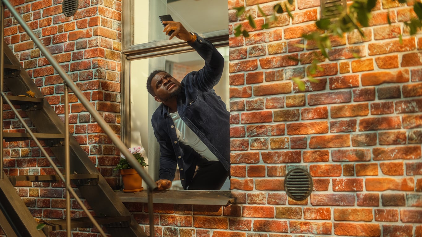 A man leans out of a window in a brick building, reaching up with his phone as if trying to get a signal. A metal staircase is next to him, and a potted plant sits on the windowsill.
