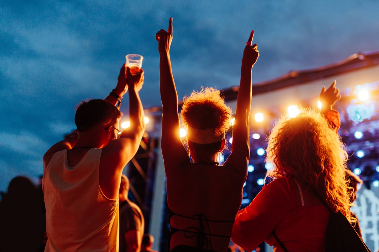 Three people at an outdoor concert at dusk, facing a brightly lit stage. Two have their hands raised, one holding a drink. The sky is darkening, creating a festive, energetic atmosphere—perfect for sharing favorite concert ticket hacks.