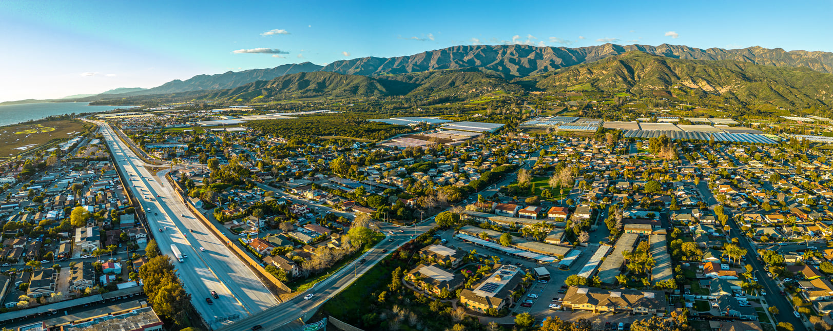 Aerial view of a suburban neighborhood with houses, tree-lined streets, a highway, and distant mountains under a clear blue sky. The coastline and ocean are visible to the left.