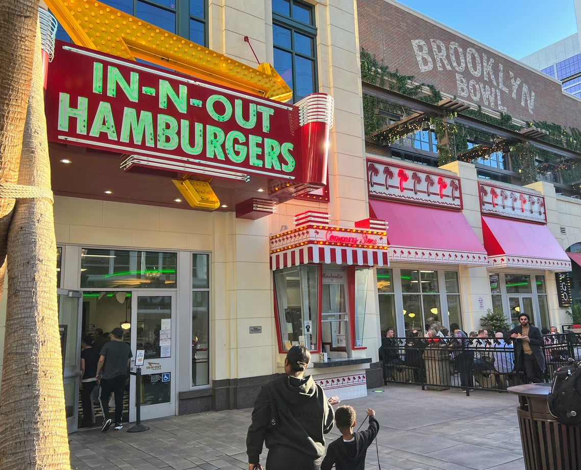People walk toward an In-N-Out Hamburgers restaurant with a retro neon sign. Next door, people dine outside at another spot. The lively urban scene showcases a classic Las Vegas budget tip: enjoy great food without breaking the bank.