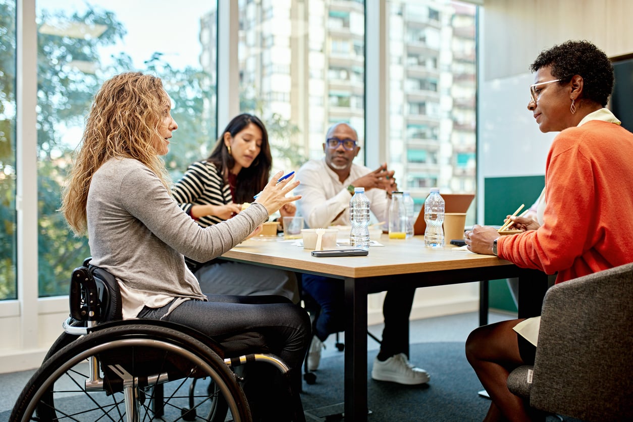 Four people are having a discussion around a table in a modern office. One woman uses a wheelchair. There are papers, water bottles, and notebooks on the table, and large windows show buildings outside.