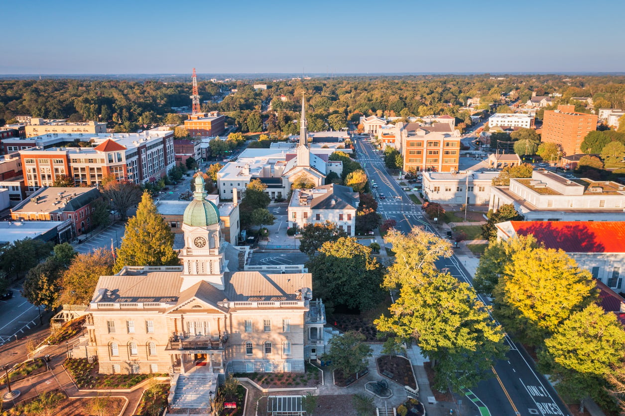 Aerial view of a historic downtown area in one of the nation’s affordable college towns, with a prominent courthouse featuring a clock tower, surrounded by trees, streets, and brick buildings under a clear blue sky.