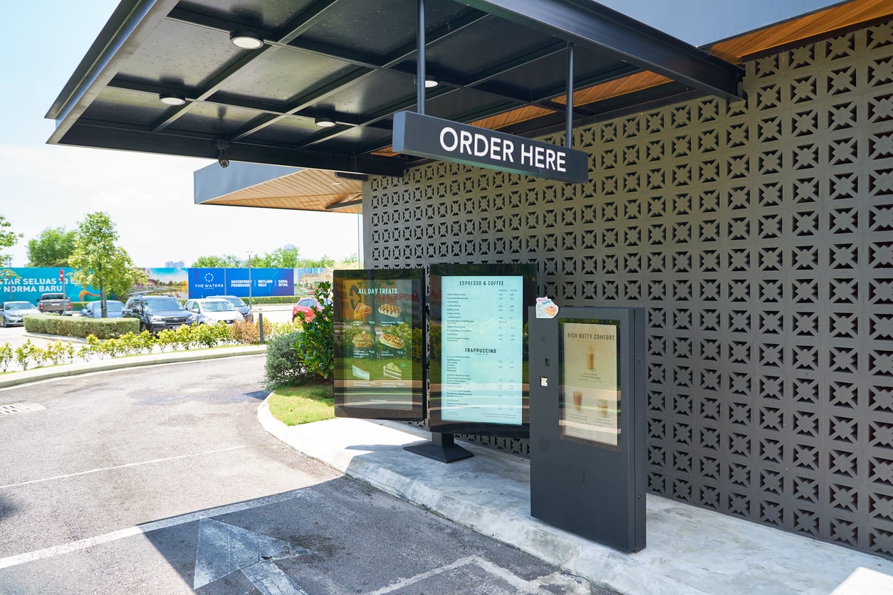 A drive-thru order station with a menu board and "Order Here" sign is next to a modern building with a patterned wall. There are trees and parked cars in the background.