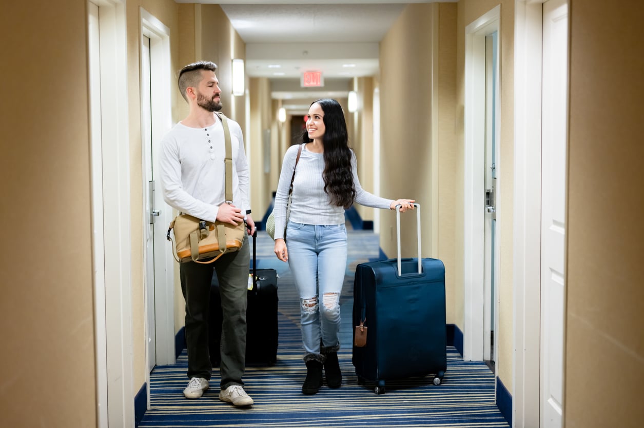 A man and woman walk down a hotel hallway with luggage, smiling at each other. Both are casually dressed and appear to be traveling together. The hall has beige walls, blue carpet, and white doors.