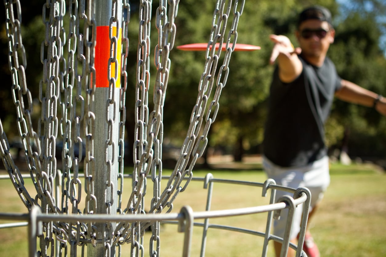 A person enjoys one of the top budget-friendly hobbies by throwing a disc towards a disc golf basket outdoors, aiming for the metal chains. Trees and grass are visible in the background on a sunny day.