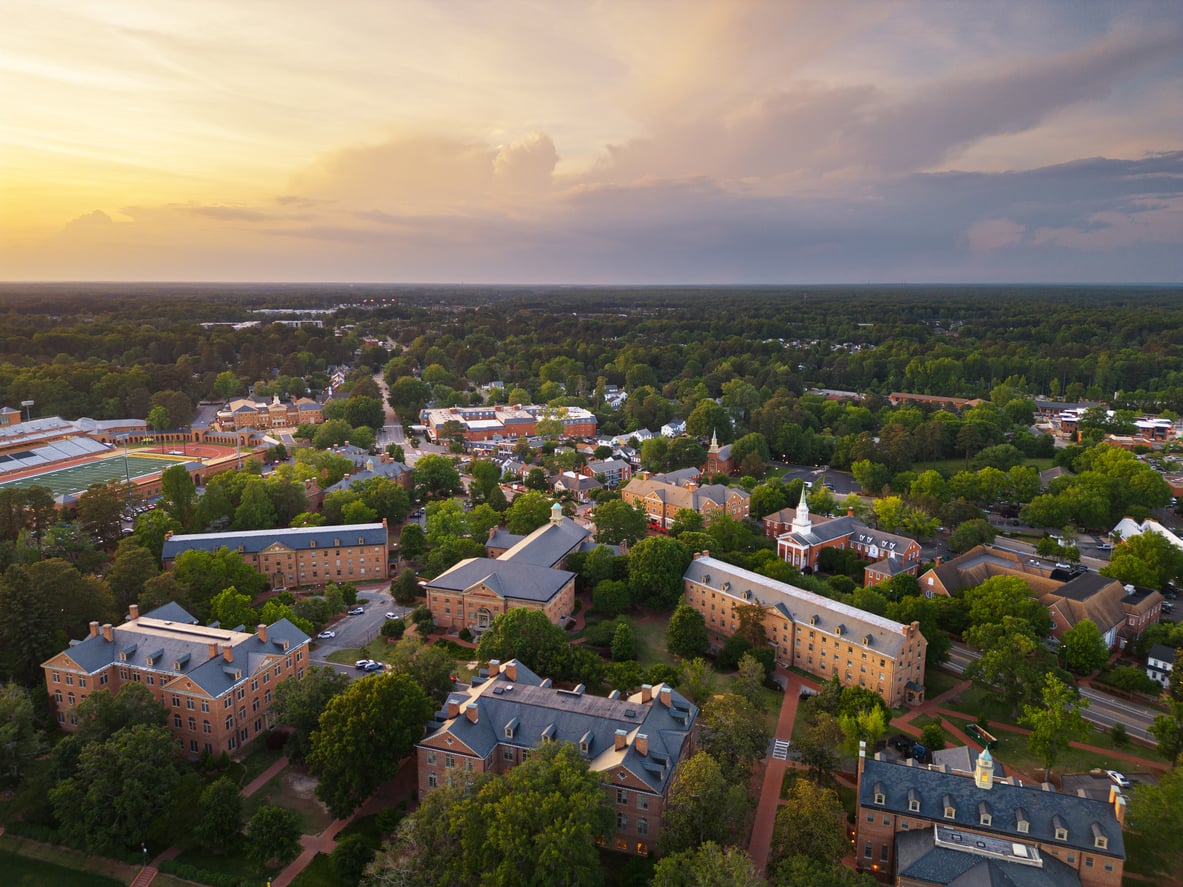 Aerial view of a college campus with historic brick buildings, green lawns, and tree-lined paths at sunset. A dome and chapel spire are visible among the buildings, surrounded by dense forest and a vibrant sky.