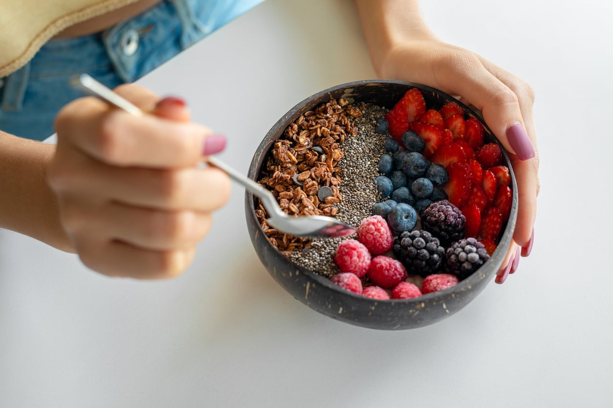 A person holding a bowl filled with granola, chia seeds (popular egg substitutes), blueberries, blackberries, raspberries, and strawberries, using a spoon to eat. The bowl is on a white surface.