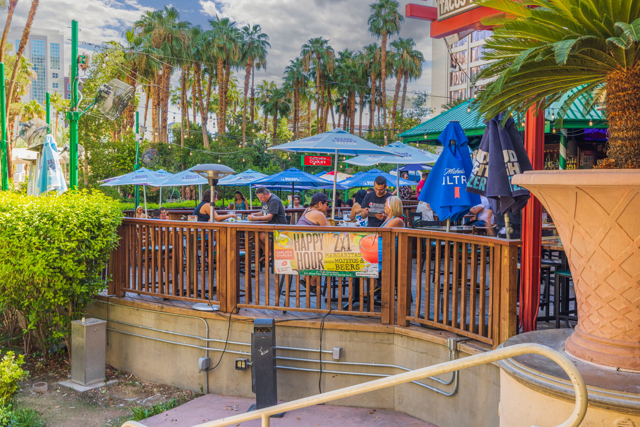 An outdoor restaurant patio with people sitting at tables under blue umbrellas, surrounded by palm trees—a perfect Las Vegas budget tips spot. A banner on the wooden railing advertises "Happy Hour" specials beneath a partly cloudy sky.