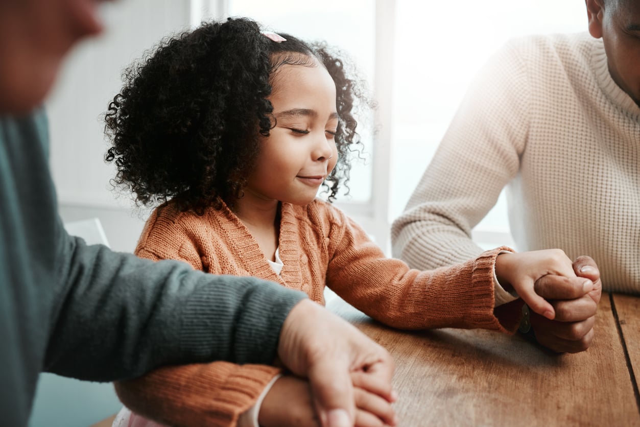 A young girl with curly hair and a brown sweater sits at a table, holding hands with two adults on either side in a gesture of prayer or support—creating a warm, peaceful atmosphere focused on connection and mental health.