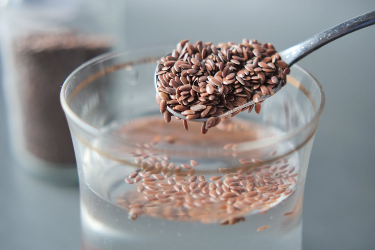 A close-up of a spoonful of brown flaxseeds, popular as egg substitutes, held above a glass of water with more flaxseeds floating inside. A blurred container of flaxseeds is visible in the background.