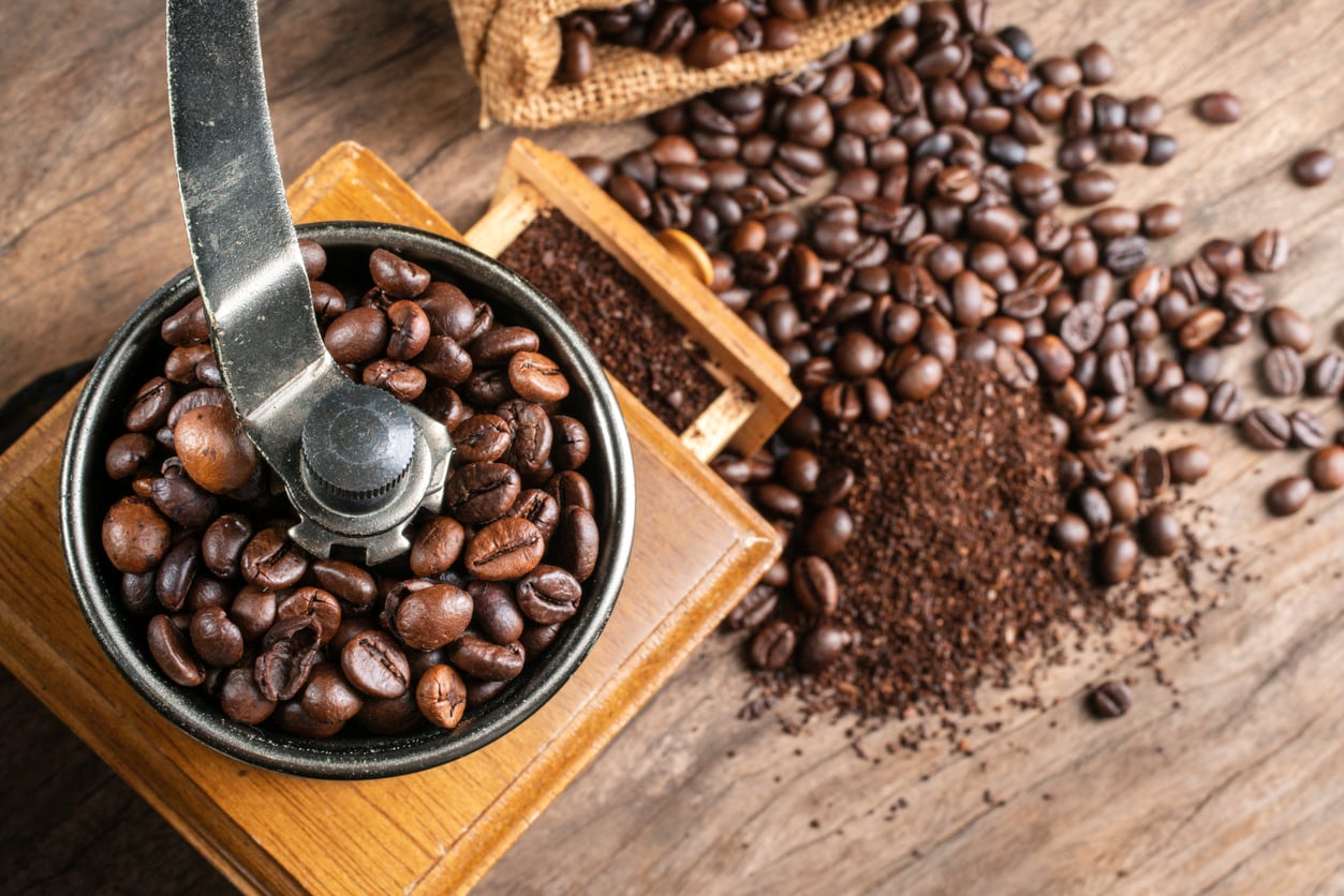 Close-up of a manual coffee grinder filled with whole coffee beans, with ground coffee and more beans spilling from a burlap sack onto a wooden surface.