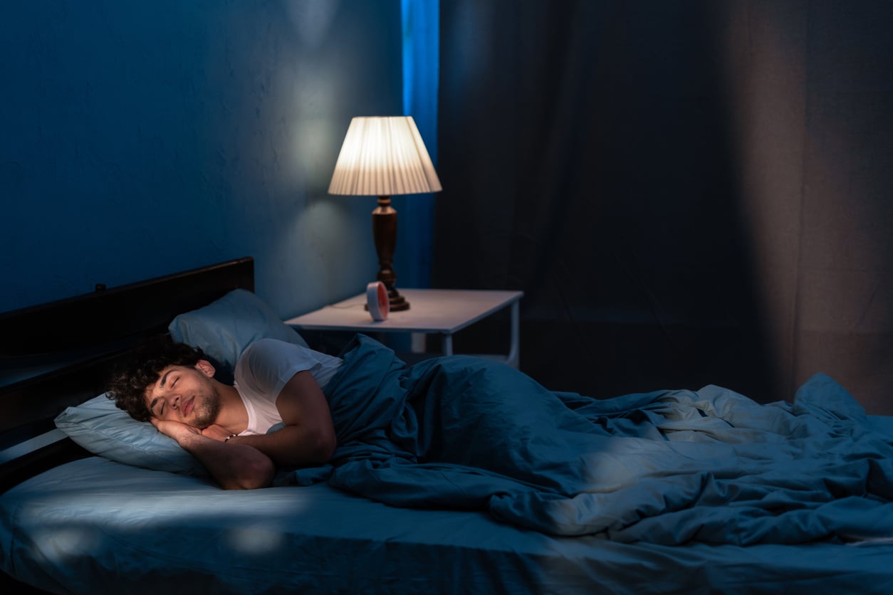 A man lies awake in bed at night under blue covers, resting on his side with a thoughtful expression—reflecting on mental health. A lit lamp and alarm clock sit on the bedside table, softly illuminating the dim room.