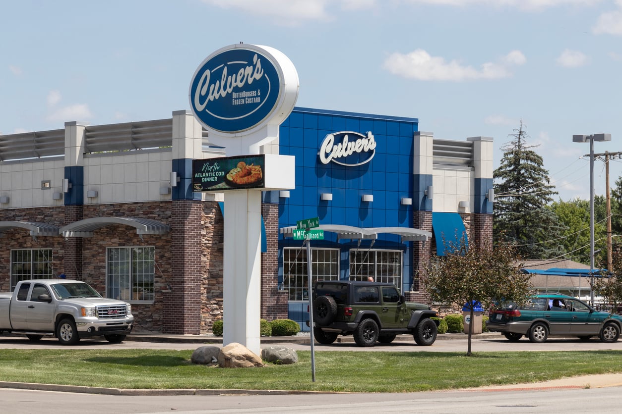 A Culver’s fast food restaurant with blue signage is shown on a sunny day. Several vehicles are parked outside, and trees and a street sign are visible in the foreground.