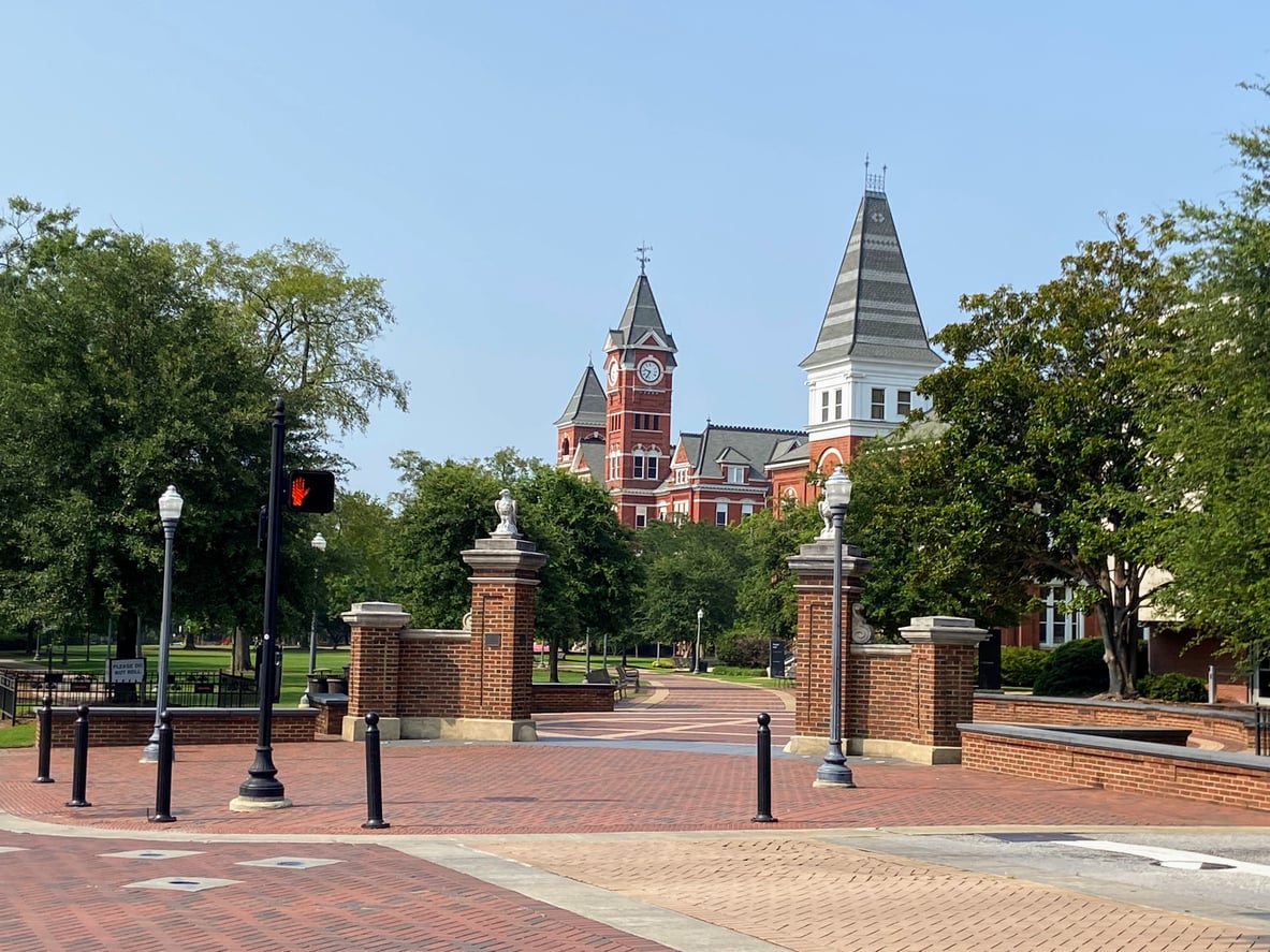 A brick-paved walkway leads to an ornate brick entrance with stone pillars, surrounded by trees—capturing the inviting charm often found in affordable college towns. In the background, a tall red brick building with a clock tower rises under a clear blue sky.