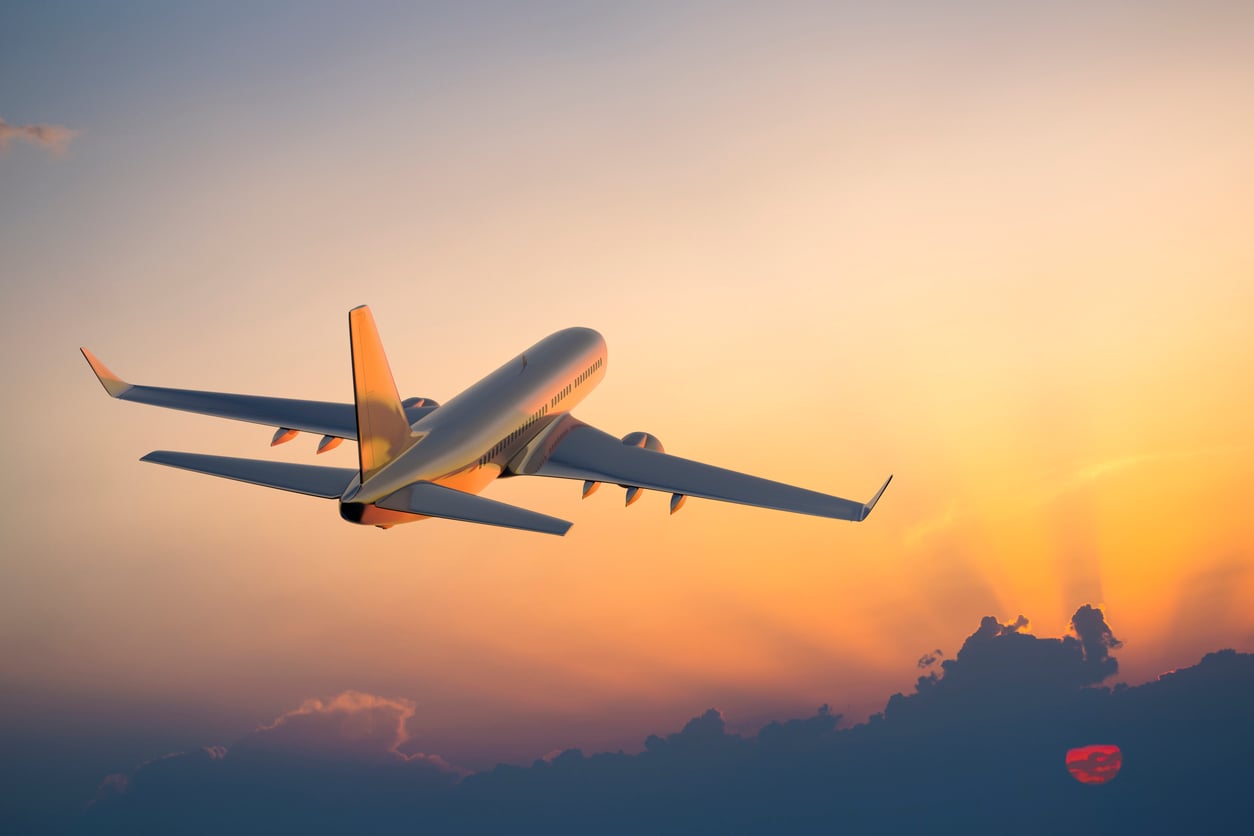 A commercial airplane flies upward into a colorful sunset sky, with sunlight illuminating the clouds and the aircraft reflecting warm orange hues.