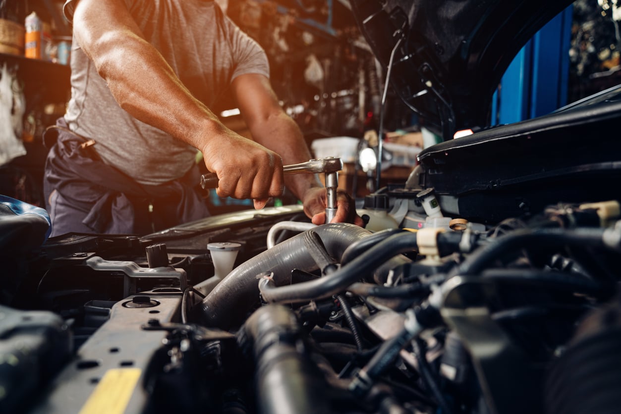 A mechanic with strong DIY skills uses a wrench under the hood of a car, working on the engine in a garage. The mechanic's arms are visible, and various engine components can be seen in the foreground.
