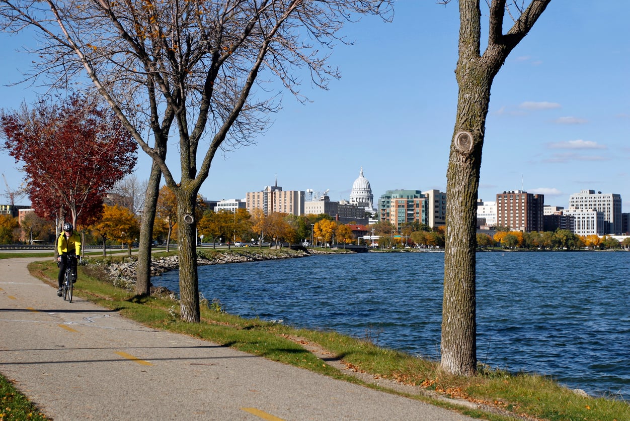 A person rides a bicycle on a lakeside path lined with trees in one of America's affordable college towns, with a city skyline and a white-domed capitol building visible in the background under a clear blue sky.