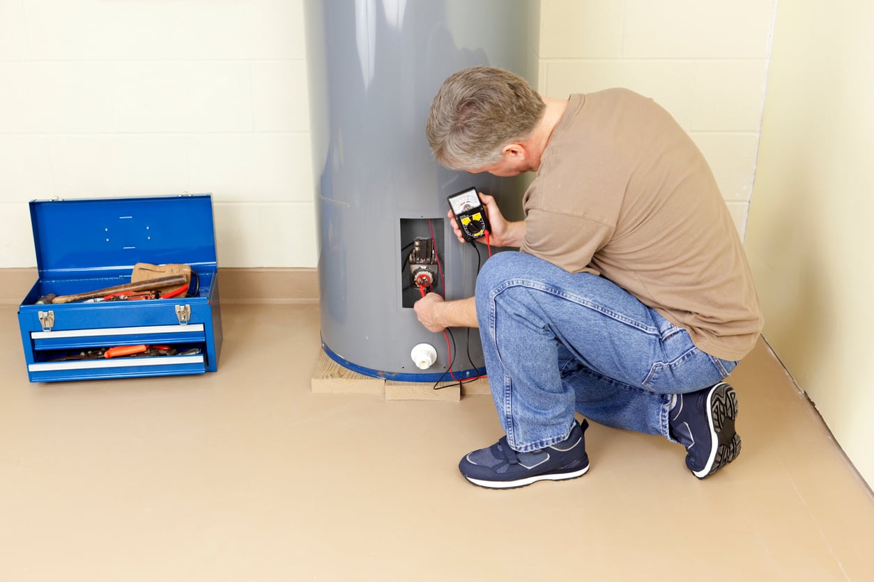 A man with strong DIY skills kneels on the floor, testing electrical components inside a water heater with a multimeter. A blue toolbox filled with tools sits open nearby. The scene unfolds indoors against a light-colored wall.
