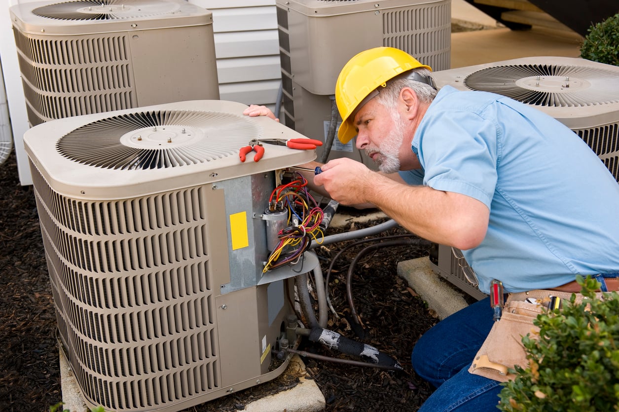 An HVAC technician wearing a yellow hard hat and tool belt is repairing an outdoor air conditioning unit, addressing issues from delayed purchases, using a screwdriver to work on exposed wiring. Several similar units are nearby.