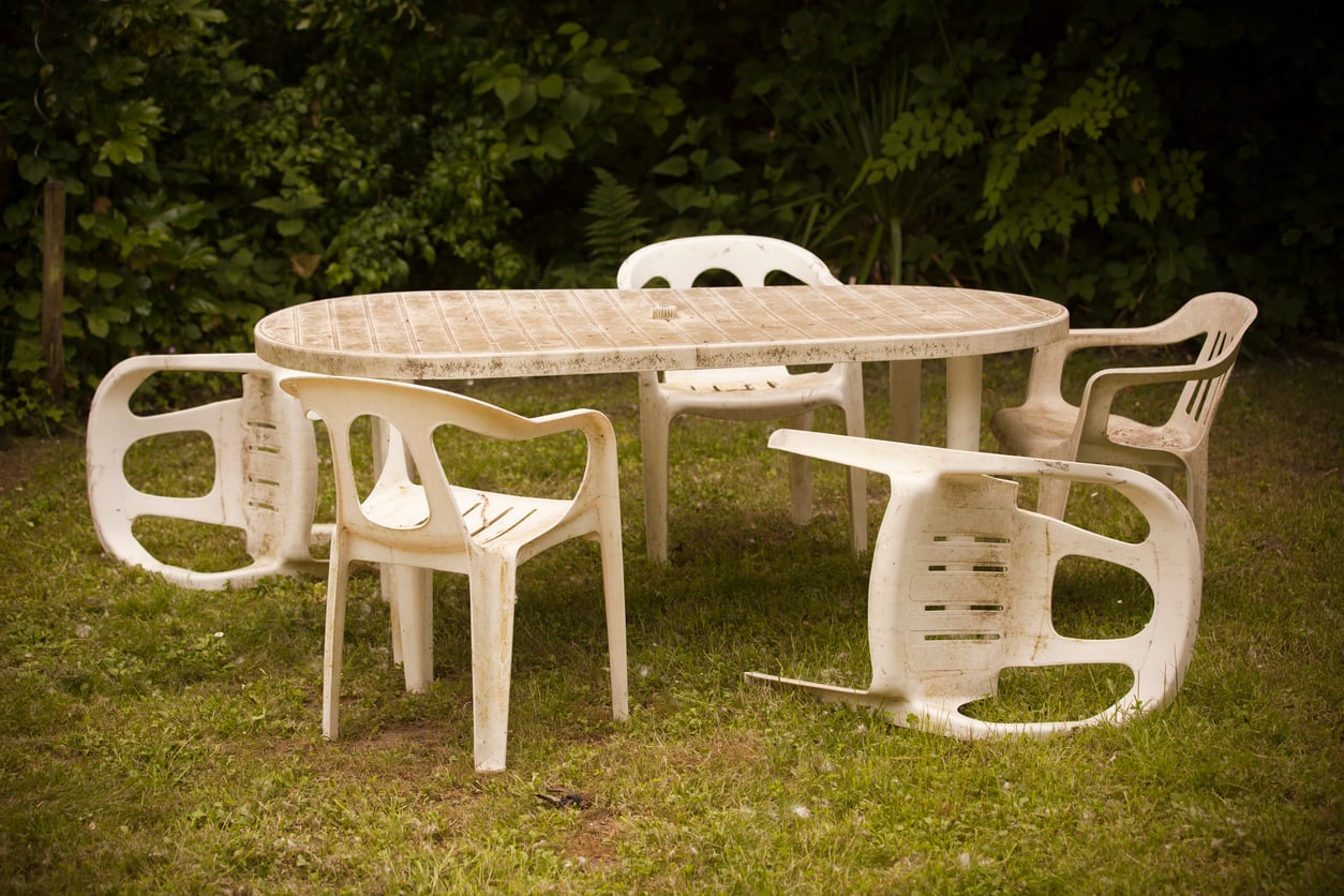 An outdoor oval table surrounded by five white plastic chairs on grass; one chair is tipped over. Lush green bushes fill the background. The furniture appears weathered and dirty.