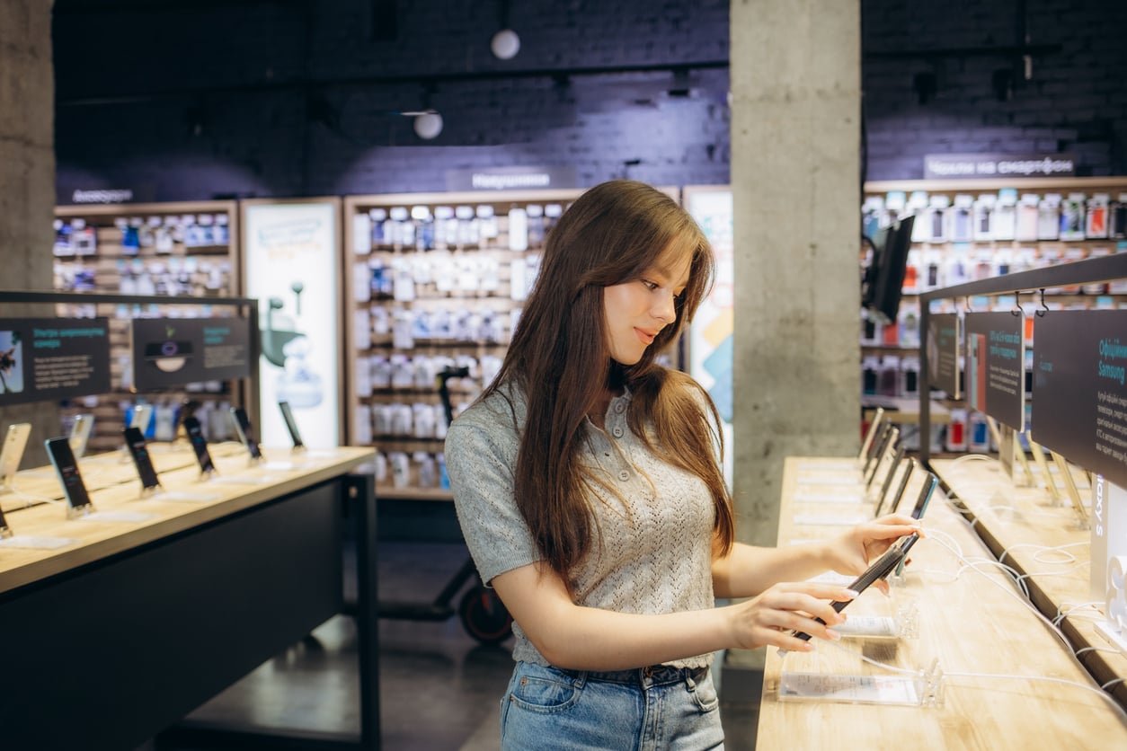 A woman with long brown hair examines a smartphone on display in a modern electronics store, surrounded by other phones and tech accessories on wooden tables.