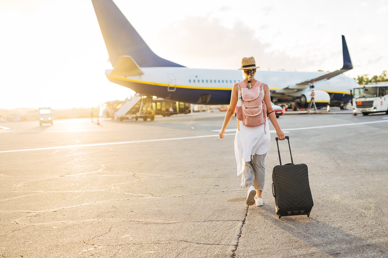 A woman wearing a hat and backpack walks toward an airplane on the tarmac, pulling a black suitcase behind her during sunrise or sunset.
