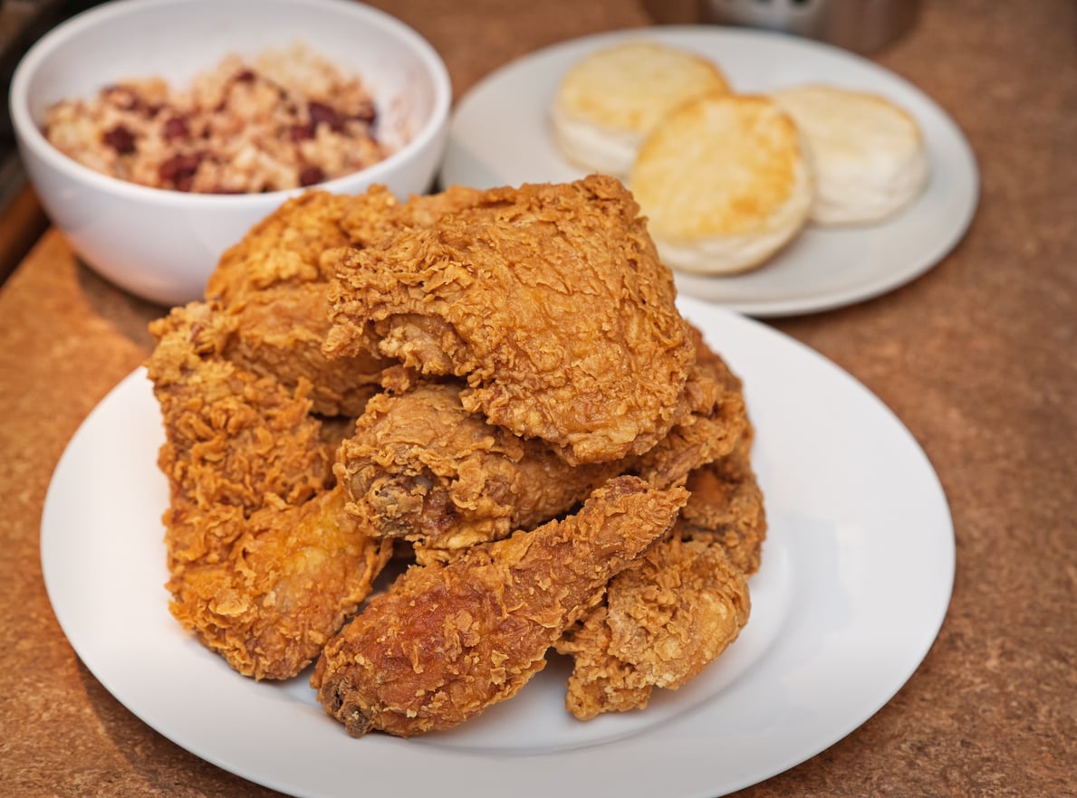 A plate of crispy fried chicken pieces sits in the foreground, with a small plate of two biscuits and a bowl of oatmeal with fruit in the background on a brown countertop.