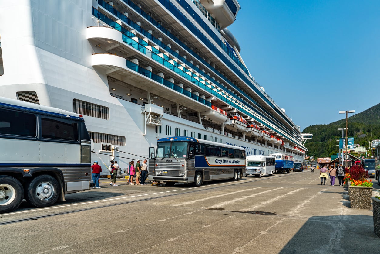 Several tour buses are parked alongside a large cruise ship at a dock, with passengers—likely eager for travel hacks—walking nearby and forested hills visible in the background under a clear blue sky.
